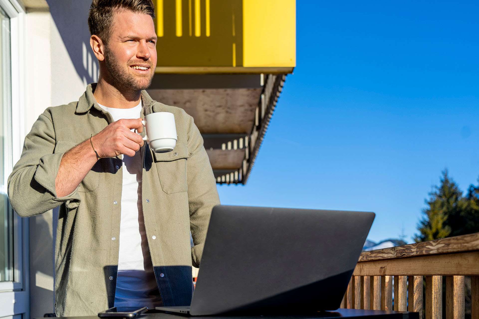 Mann mit Kaffee an einem sonnigen Wintertag auf dem Balkon mit Laptop