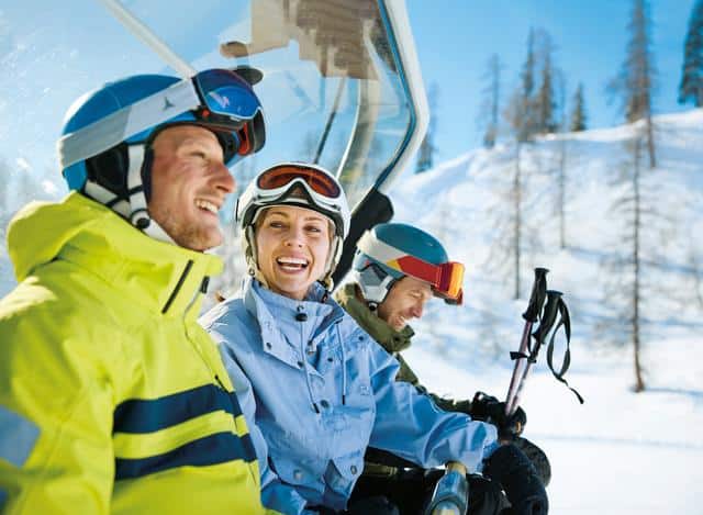 3 Personen die bei Sonnenschein im Skilift sitzen und sich auf's Skifahren freuen